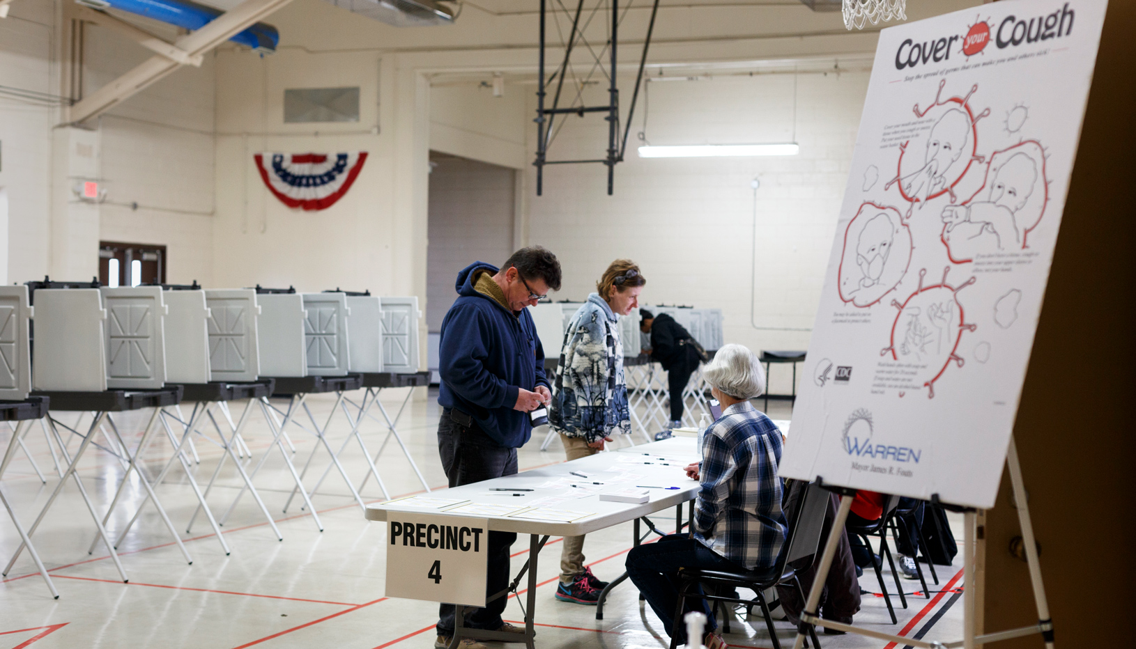 A poster with guidelines for safely coughing in the foreground of an image of a polling place.