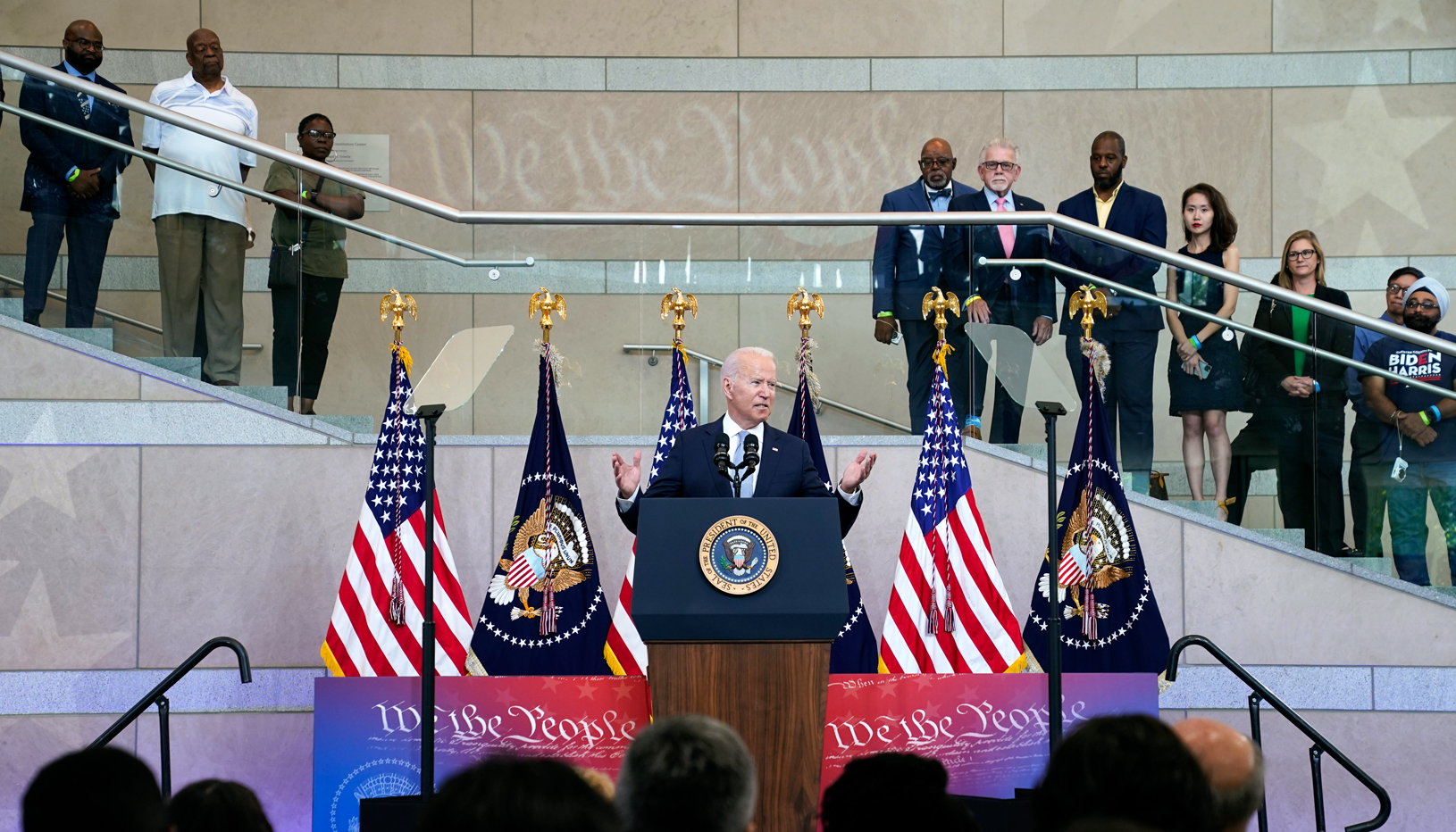 President Joe Biden speaks from a podium, surrounded by American flags and onlookers