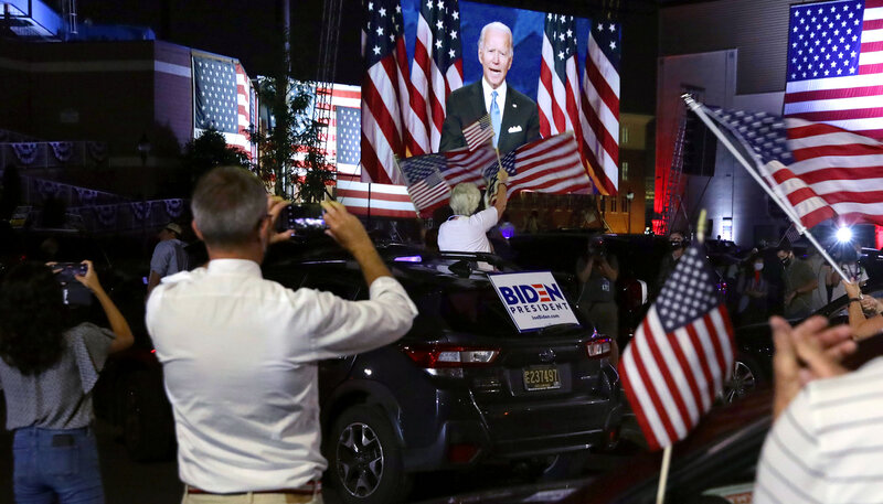 Joe Biden is shown on a screen delivering a speech to a crowd of people and cars