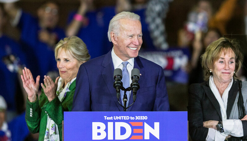 Joe Biden smiles at a crowd from a podium, while Jill Biden and Valerie Biden applaud behind him