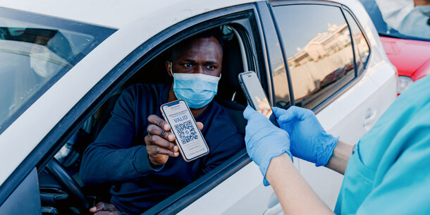 A man inside his car shows his vaccine credentials to a health official through an open window