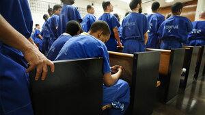 Prisoners lined up in a court house. Man is seen hunched over as he sits and waits in a county jail uniform.