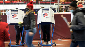 person walking past two voting booths while wearing a mask.