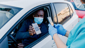 A man inside his car shows his vaccine credentials to a health official through an open window