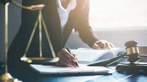 A suited woman writes on a notepad, near a gavel and scale